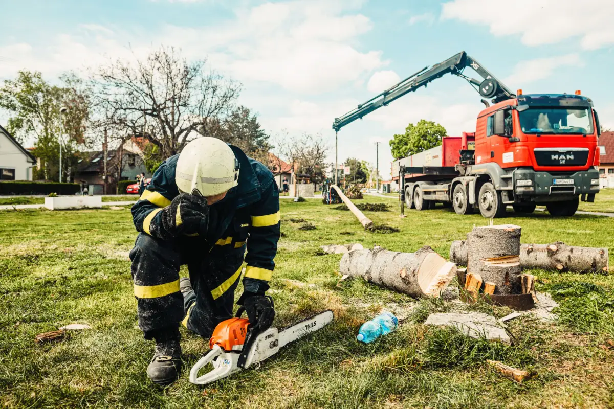 A worker in a helmet and protective gear kneeling beside a chainsaw, with cut trunk rounds and a crane truck behind