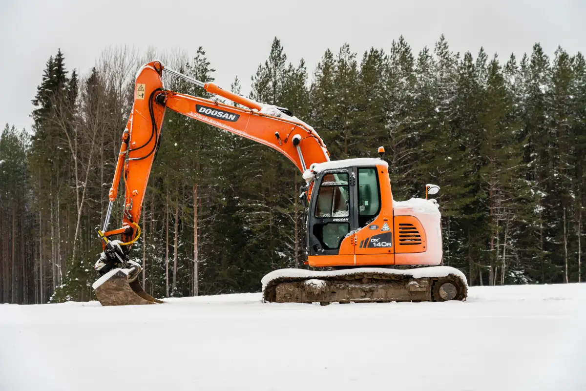 An excavator standing on snow-covered ground in front of a conifer treeline
